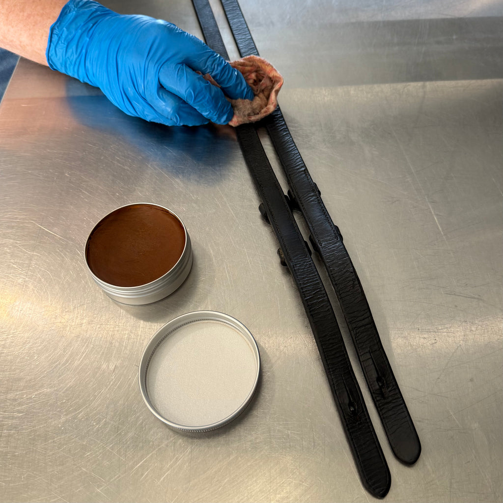 Person cleaning a leather bridle with 'Fire Horse Botanicals' balm and cloth on a metal surface.