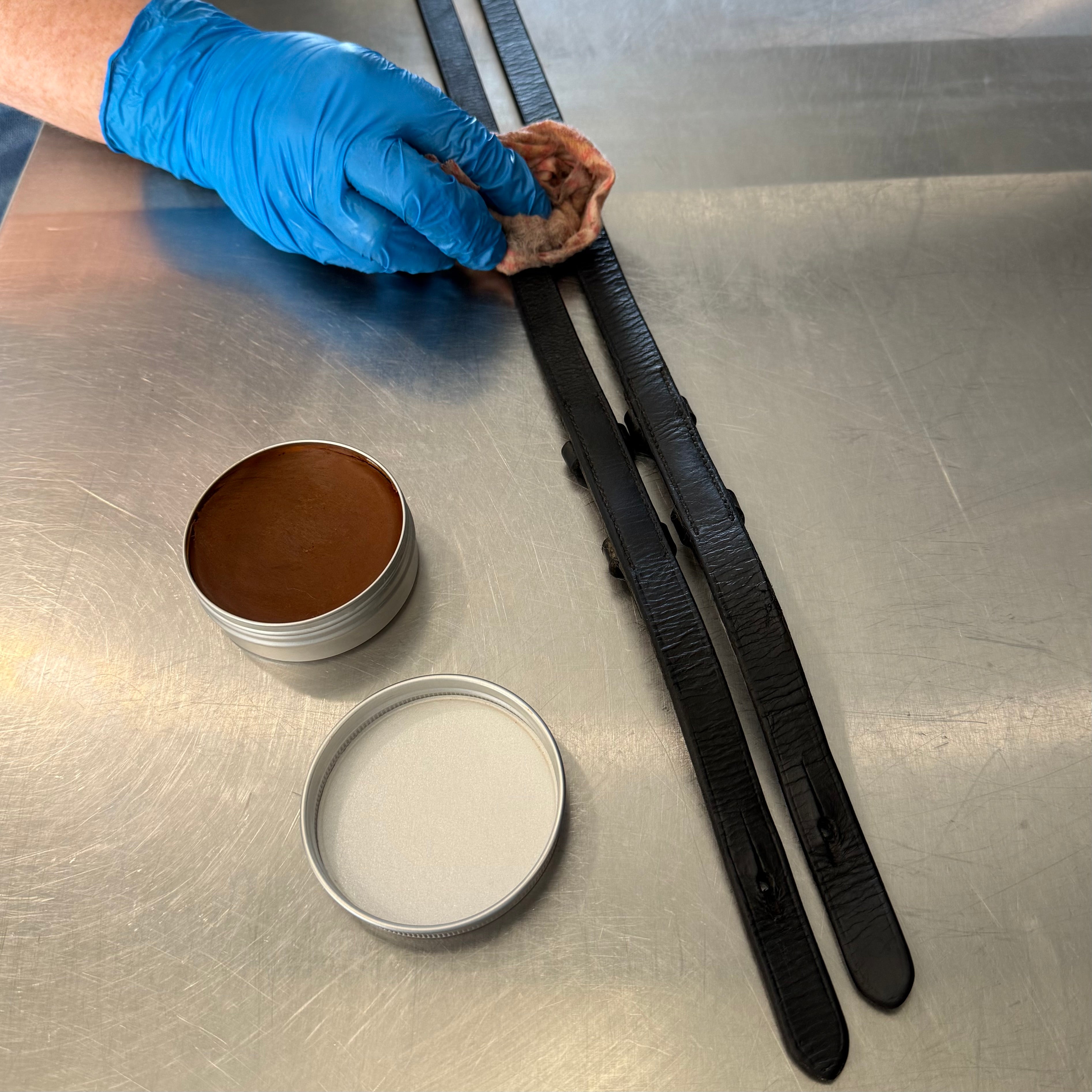 Person cleaning a leather bridle with 'Fire Horse Botanicals' balm and cloth on a metal surface.