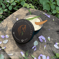 Tin of 'Fire Horse Botanicals' gel with a wooden applicator on a wooden surface with flowers and plants in the background
