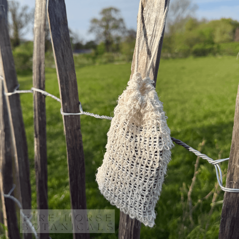 White sisal soap bag hanging on a wooden fence in a grassy field with 'Fire Horse Botanicals' branding