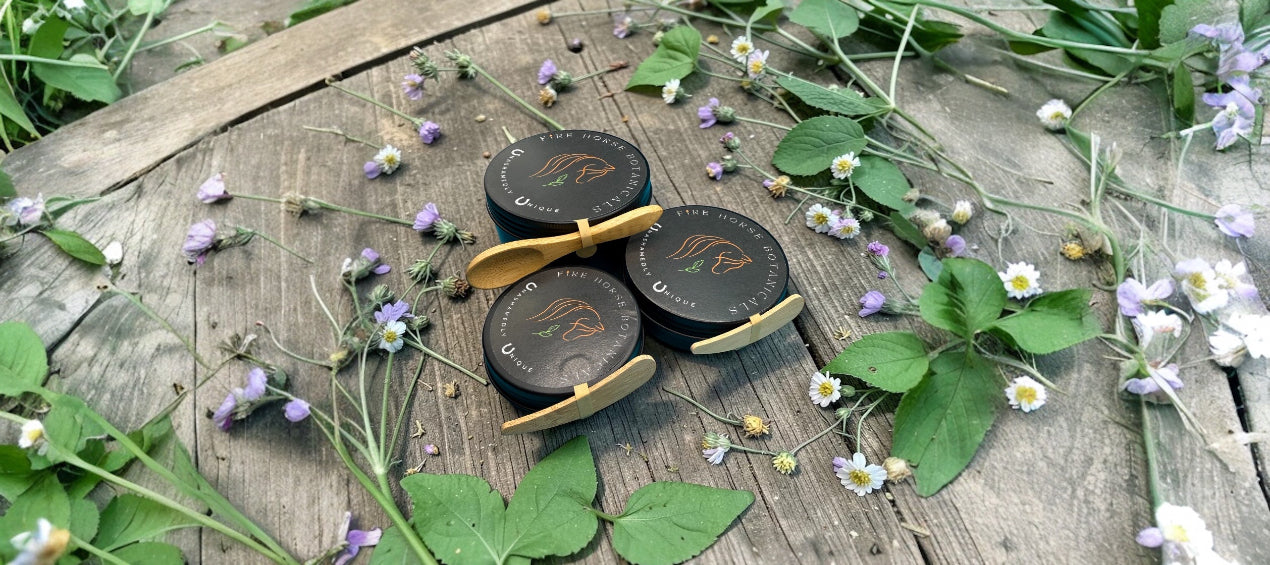 Three Fire Horse Botanicals tins on a wooden surface with flowers and leaves.
